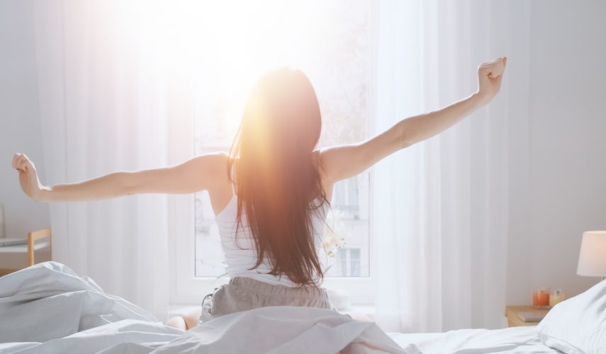 Beautiful Brunette is Waking up in the Morning, Stretches in the Bed, Sun Shines on Her From the Big Window. Happy Young Girl Greets New Day with Warm Sunlight Flare